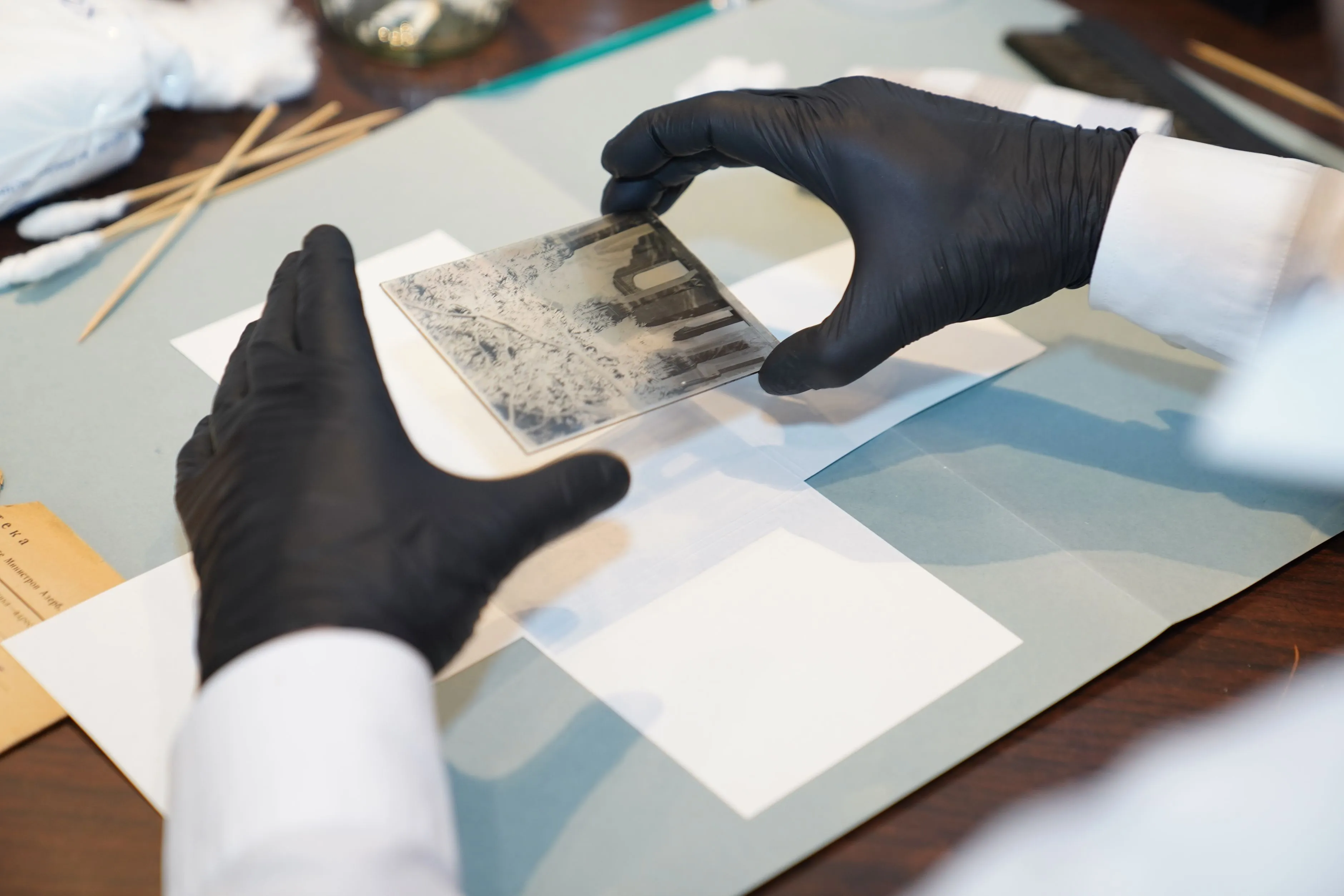 Gloved hands holding a small black and white photograph over archival tools on a workbench