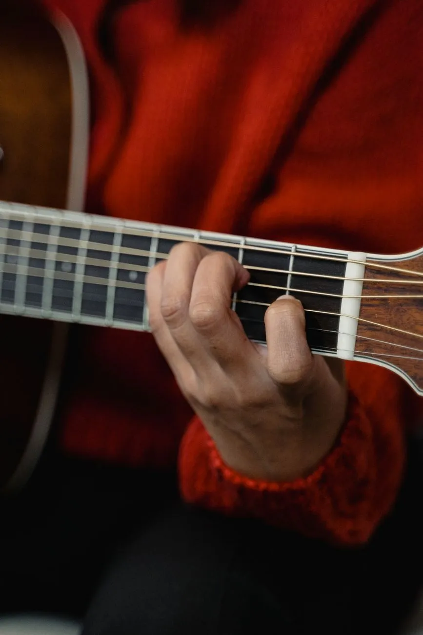 A hand in a red sweater pressing down on the fretboard of an acoustic guitar, fingers curled around the neck