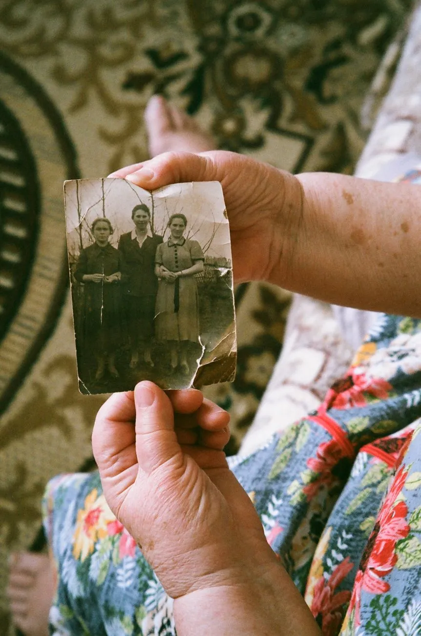 Weathered hands holding a small, cracked and torn sepia photograph of three women standing outdoors