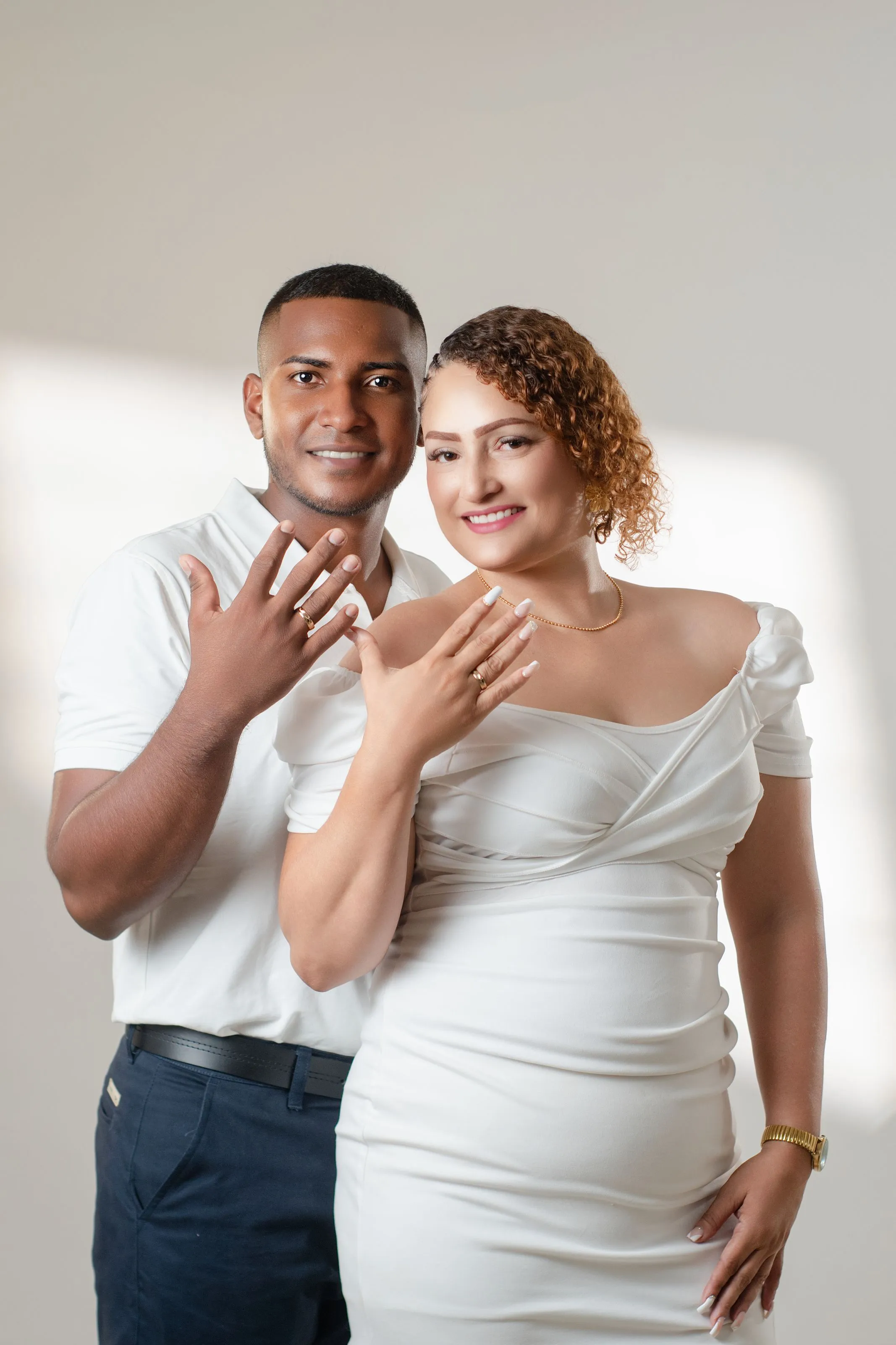 A couple in white smiling closely together indoors in soft, even light, both hands raised showing their rings